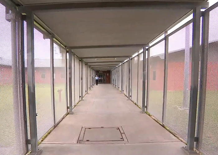 Long outdoor corridor inside a prison with mesh fencing and a guard walking ahead