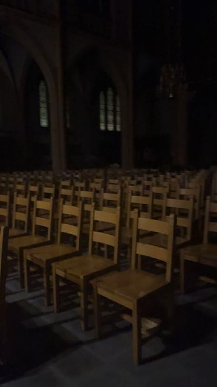 Rows of empty wooden chairs in a dark, cavernous hall, creating a haunting liminal space with deep shadows.