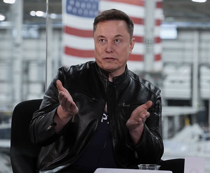 Elon Musk speaking during an interview with American flag in background