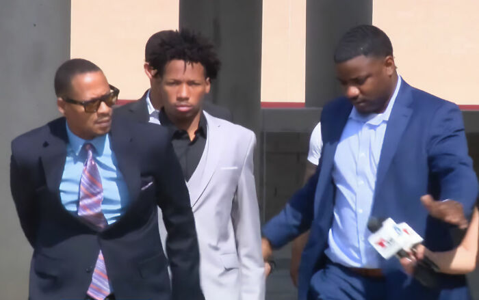 Group of men with one in handcuffs leaving building with media present
