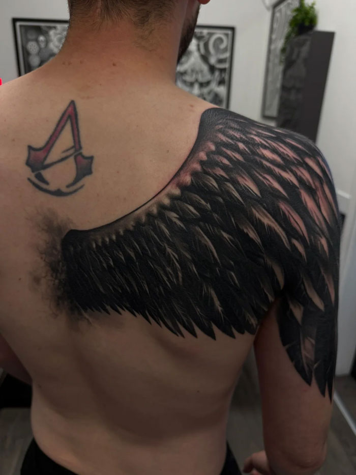 A tattoo artist's work featuring a large black wing and the Assassin's Creed logo on a person's back, a questionable tattoo idea.