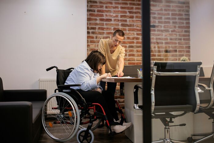 Person assisting wheelchair user at table in modern office environment