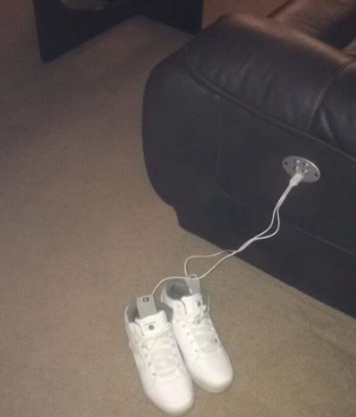 A pair of white sneakers plugged into an electrical outlet on the side of a black leather couch, a unique interior designer find.
