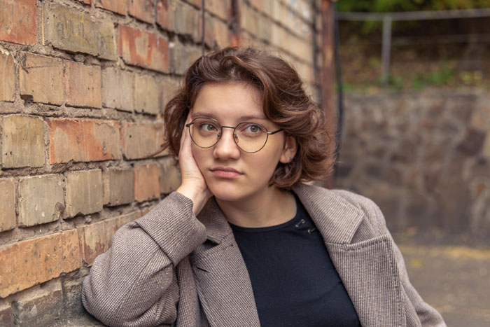 A young woman with short brown hair and round glasses leans against a brick wall, appearing thoughtful or worried about a toxic guy.