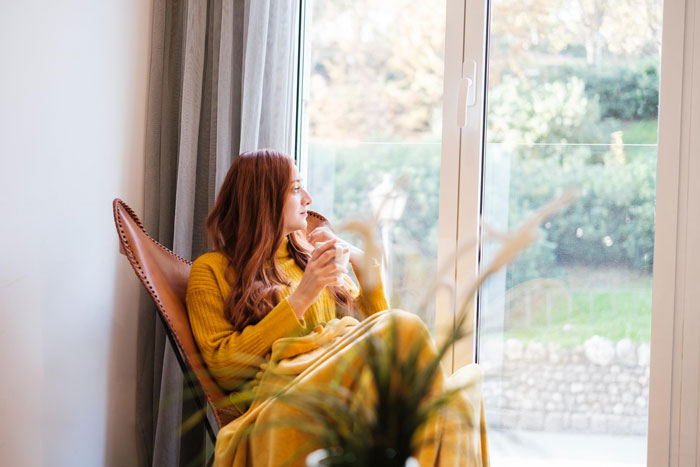 A woman sits by a window, covered in a yellow blanket, looking out, enjoying peace after a toxic relationship.