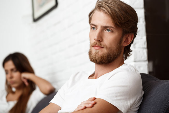 A toxic guy with a beard and white shirt, arms crossed, looking away from his girlfriend, showcasing his jealousy.