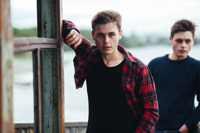 A toxic guy looking at the camera, leaning against a metal structure with another guy blurred in the background, highlighting the jealousy and rules.