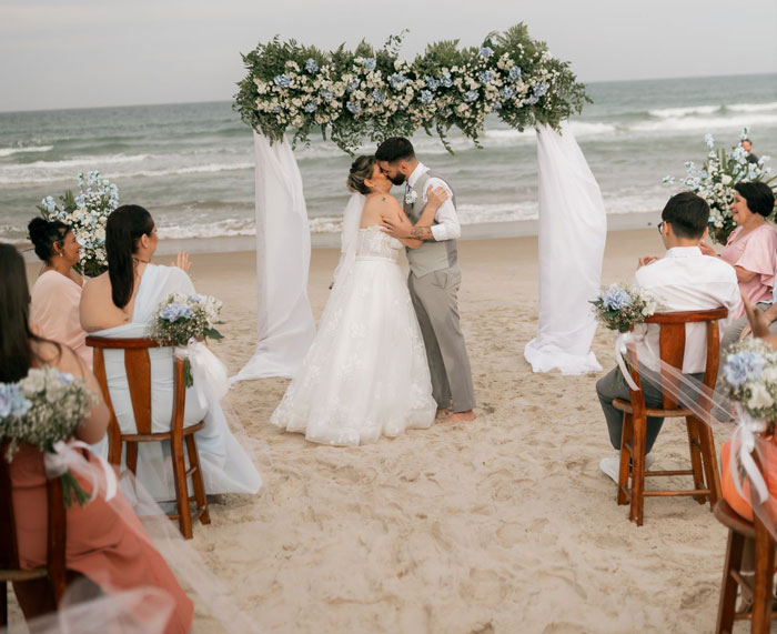 Couple kissing at beach destination wedding ceremony under flower arch