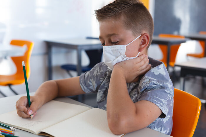 A young student wearing a mask and writing in a notebook in a classroom, showing students learning from teachers.