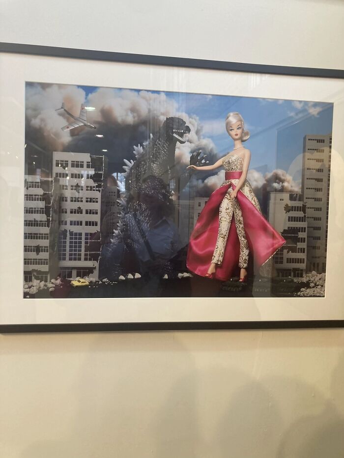 A framed photograph depicting a giant Godzilla and a Barbie doll in a city, a bizarre image.