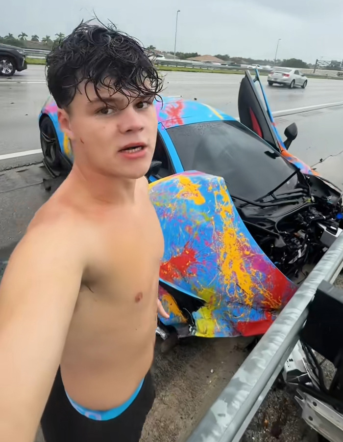 A young man stands next to his heavily damaged, colorful sports car on a highway, part of the Celebrity GoFundMe Scandals.