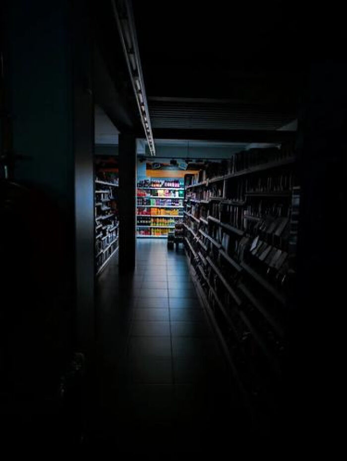 A dark, haunting liminal space in a library aisle with dimly lit colorful shelves at the end.