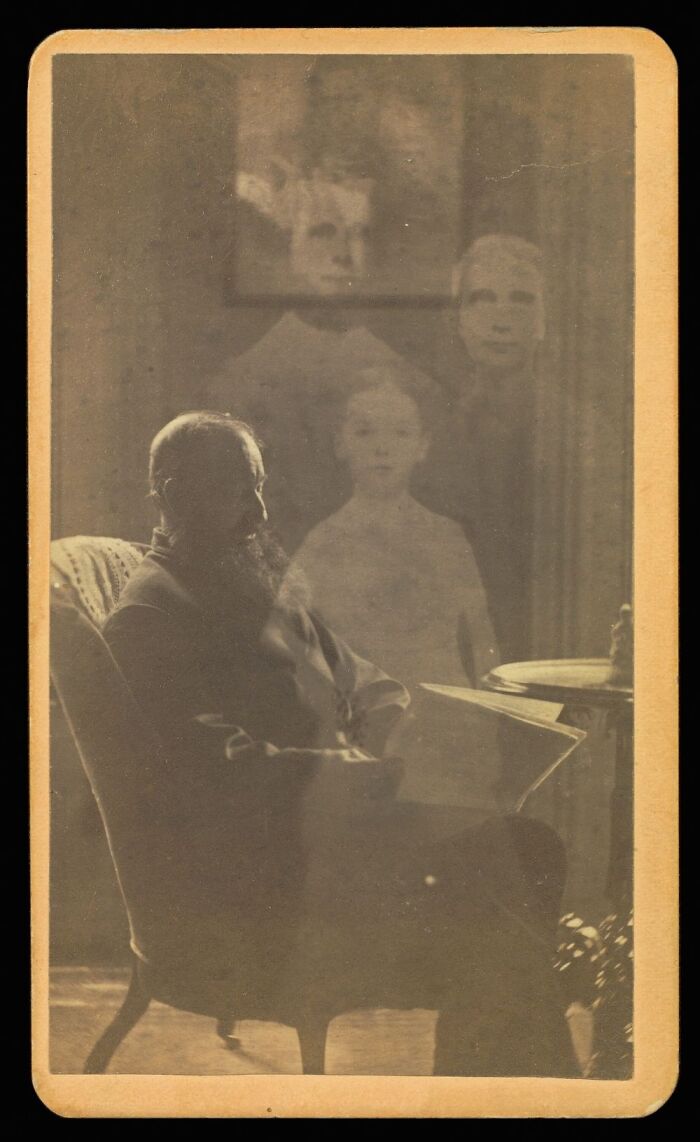 An eerie historical photo of a man seated with several ghost-like figures of children appearing behind him.