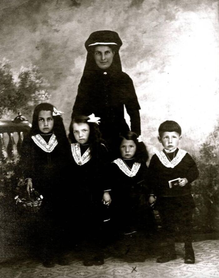 A somber historical photo of a woman in dark attire standing with four young children, all in dark clothing.