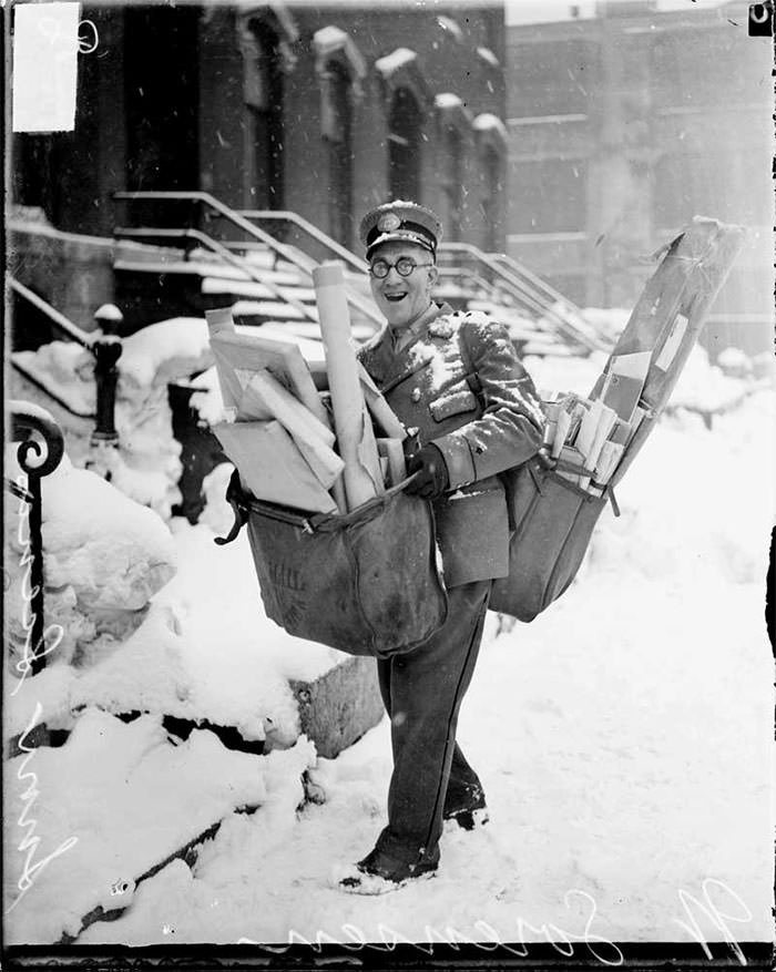 Historical photo: a smiling mailman delivering an enormous amount of mail, walking through a snowy landscape.