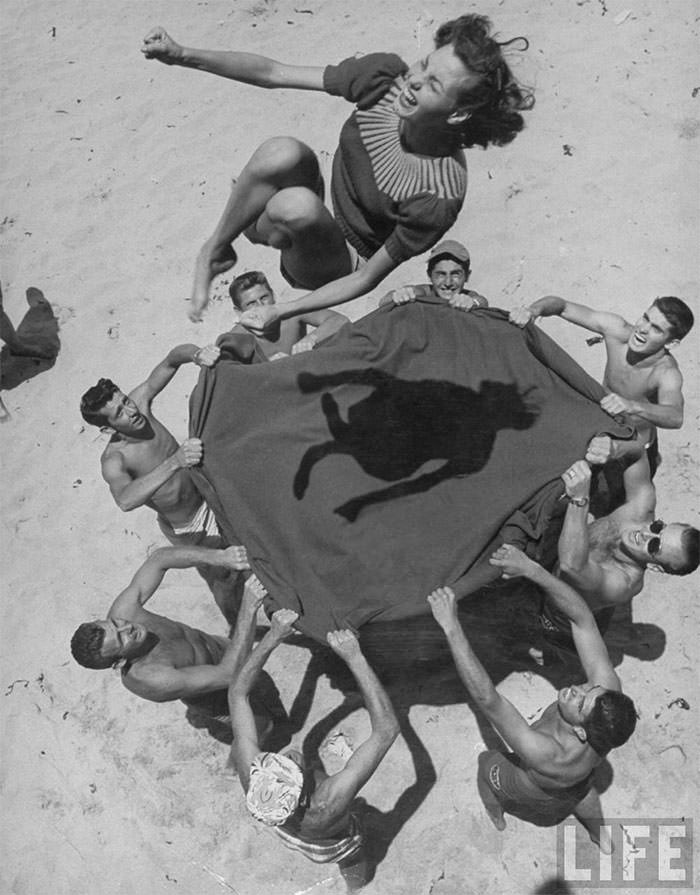 Historical photo: a woman is being tossed in the air by a group of men on a beach, creating a memorable shadow on the blanket.