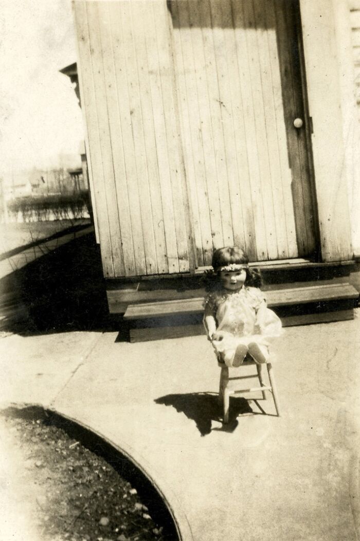 A small child in a flower crown sits on a stool outside a wooden building in a historical photo.