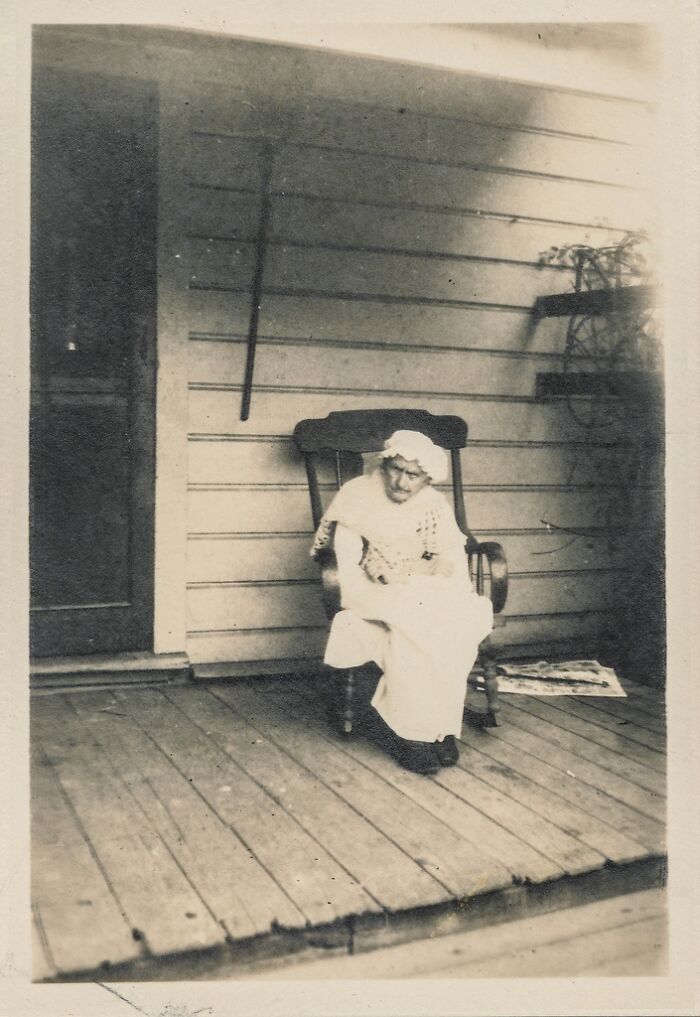 An elderly woman in a white cap sits in a rocking chair on a porch, a historical photo.