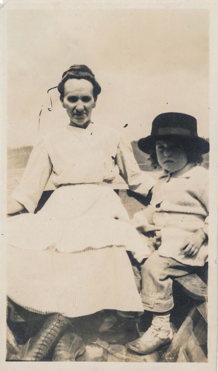 Black and white historical photo of a woman in a striped dress and a child in a hat sitting outdoors.