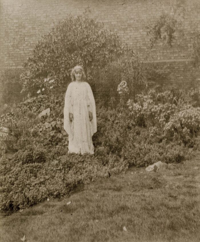 Sepia-toned historical photo of a person in a white gown standing in a overgrown garden.