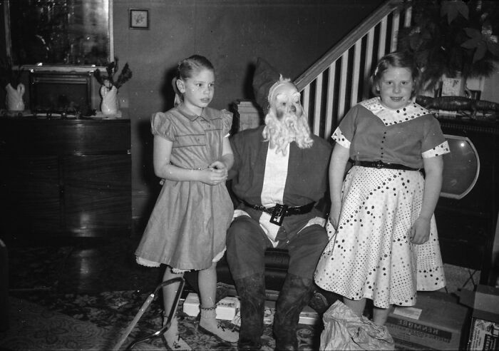 Black and white historical photo of two young girls posing next to a Santa Claus figure indoors.