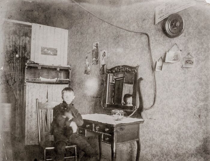 Sepia historical photo of a boy holding a cat while sitting at a dressing table in a patterned room.