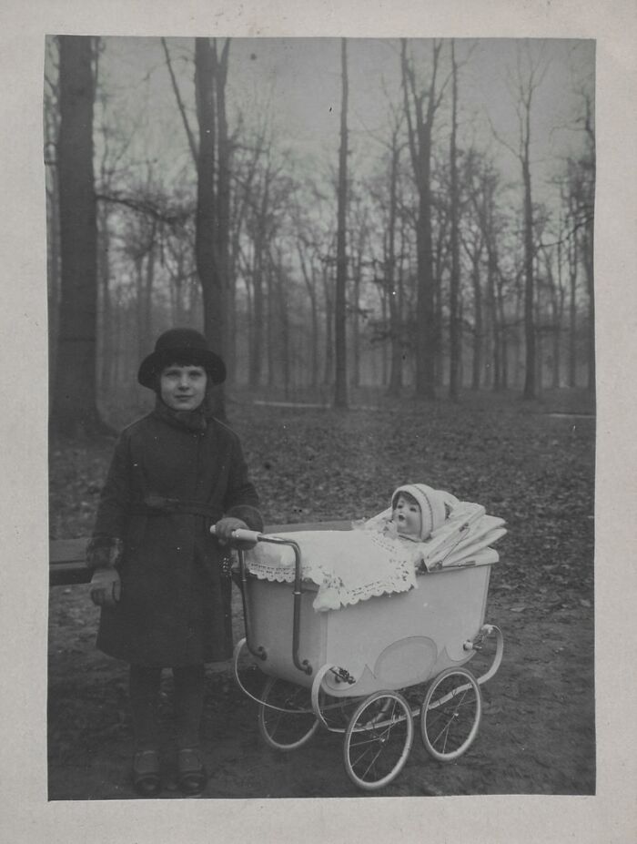 A chilling historical photo of a child standing next to a doll in a pram, set against a backdrop of bare trees, an unexplainable image.