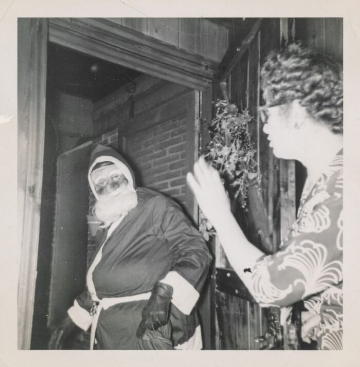 Black and white photo of Santa entering a doorway, a woman reaching out to him. Historical photo.