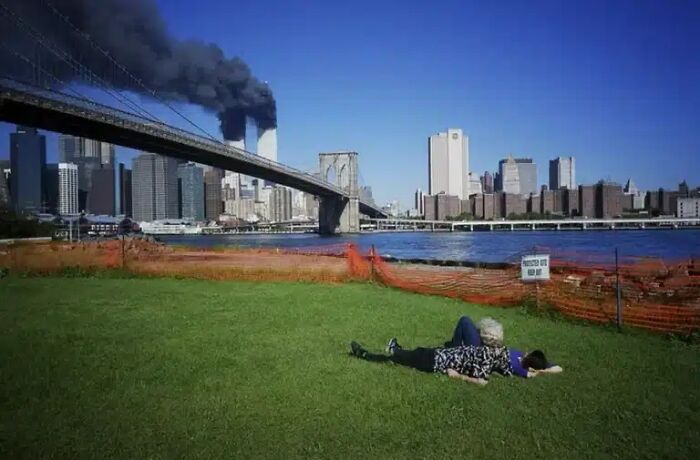 Two people relaxing on grass by a river with the Twin Towers burning in the background, one of many historical photos.