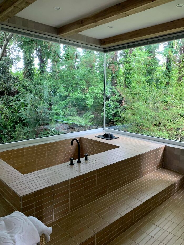 Modern bathroom with large brown tiled sunken bathtub and corner windows showing greenery