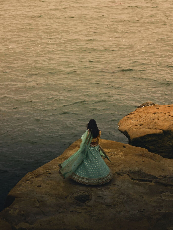 Aesthetic woman in flowing dress standing on rocky shore by calm water