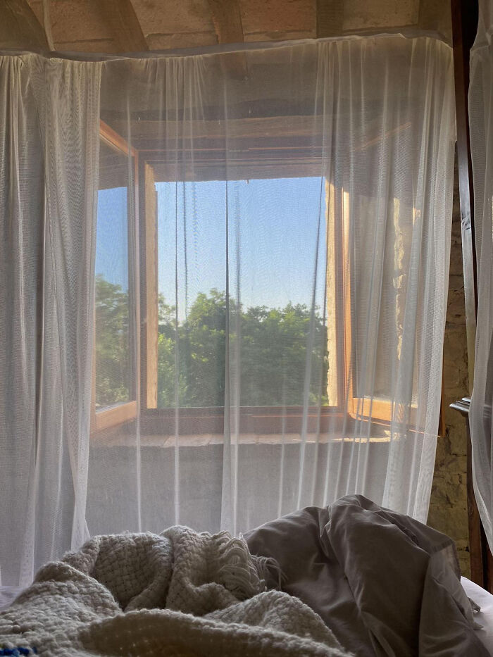 Cozy bed with knitted blanket by window framed with sheer curtains and greenery outside