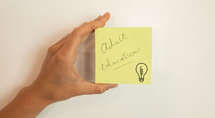 A hand holds a yellow sticky note with Adult Education and a lightbulb drawing, symbolizing life skills and adulting IQ. A hand holds a yellow sticky note with Adult Education and a lightbulb drawing, symbolizing life skills and adulting IQ.
