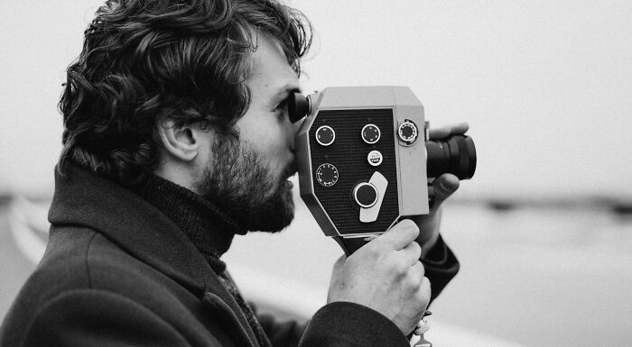 Black and white image of a bearded man looking into the viewfinder of a vintage movie camera, testing his movie memory.