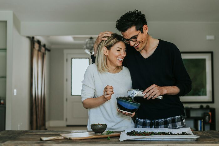 A happy couple cooking together, showing women who marry their partner. The woman is holding a bowl while the man embraces her.
