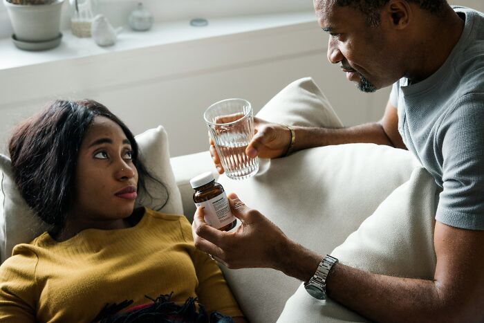 A man hands a glass of water and medication to a woman lying down, a caring moment that may lead her to marry her partner.