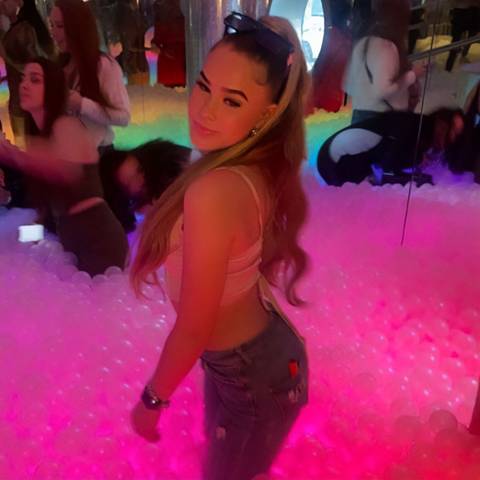 Smiling influencer in a pink top and jeans, standing in a brightly lit room filled with pink balls, reflecting the grave charges.