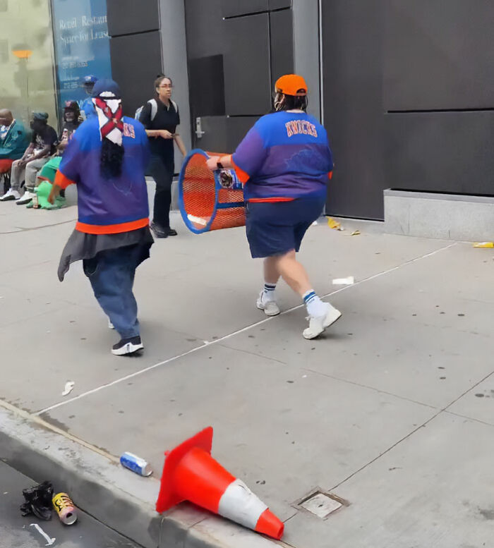 A person in a Knicks jersey walking on a street, carrying the stolen Knicks trashcan, after emptying its contents. A person in a Knicks jersey walking on a street, carrying the stolen Knicks trashcan, after emptying its contents.
