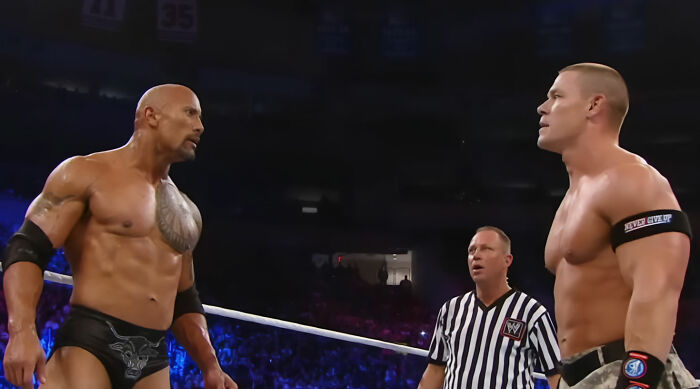John Cena in the wrestling ring facing The Rock, with a referee between them, before his trouble walking.