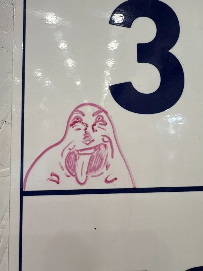 A bizarre pink cartoon face with a tongue sticking out, drawn on a whiteboard next to the number 3.