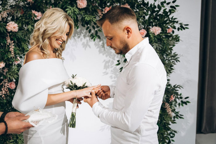 A man placing a ring on a smiling bride's finger during a wedding ceremony with floral decor.