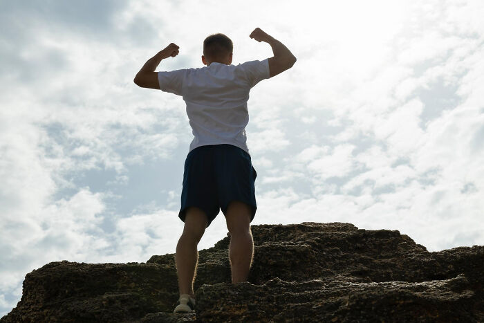 A man flexing his muscles on a rocky peak against a bright sky, representing human behavior and strength.