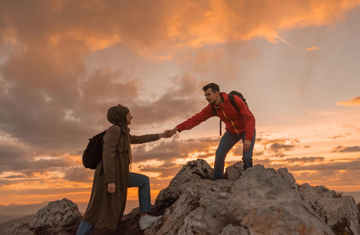 A man helps a woman climb a rocky peak, illustrating human behavior and support at sunset.