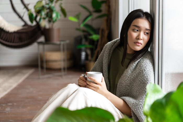 A contemplative woman by a window, holding a mug, depicting reflective human behavior.