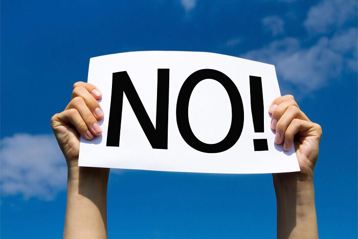 Hands holding a sign with the word NO against a blue sky, reflecting human behavior of refusal.