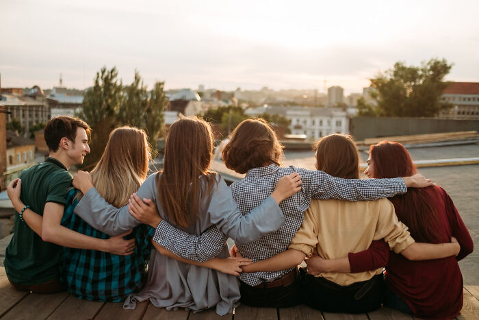 Six friends with arms around each other, sitting on a rooftop, showing human behavior and friendship.