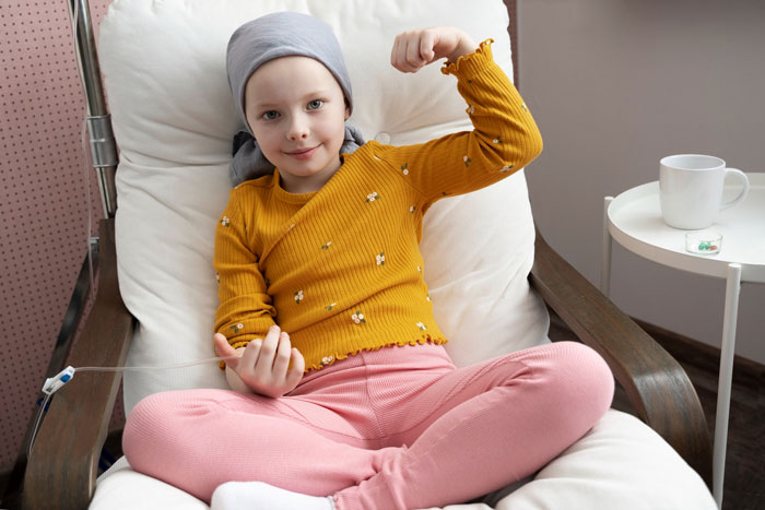 A young girl undergoing chemotherapy, wearing a head wrap and yellow shirt, flexes her arm, showing strength. A young girl undergoing chemotherapy, wearing a head wrap and yellow shirt, flexes her arm, showing strength.
