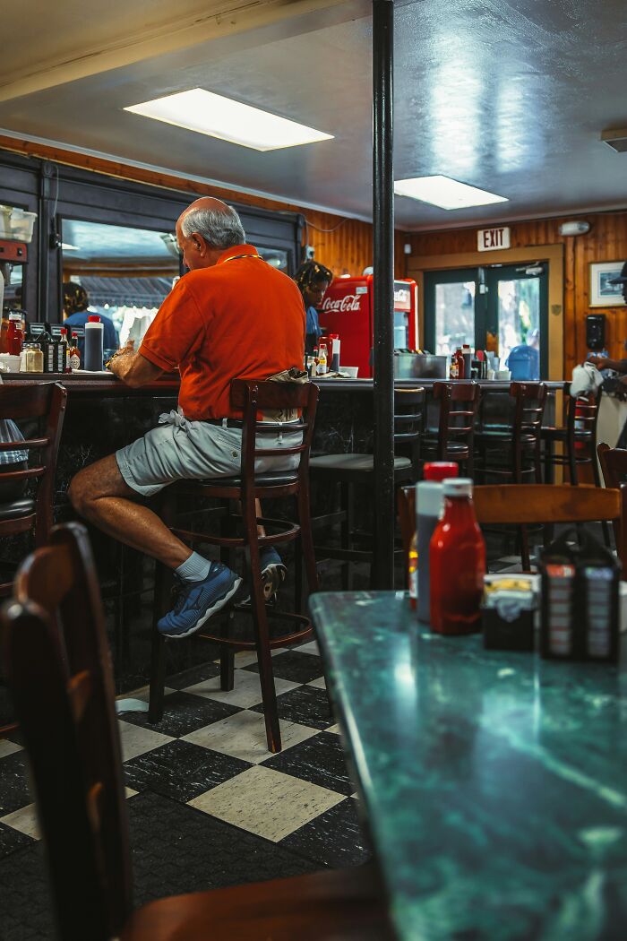 A man hides his true self at a diner, sitting at a counter in an orange shirt and shorts.