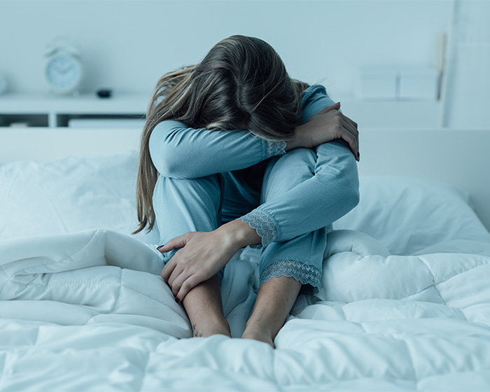 A woman sitting on her bed, visibly distressed and hugging her knees, experiencing sadness trigger syndrome.