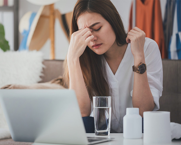 A woman experiencing sadness trigger syndrome, holding her head in distress while sitting at a desk with a laptop and a glass of water.